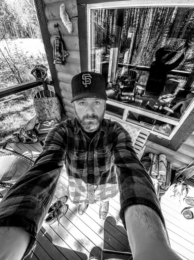 Travis on his cabin porch, arms out, taking a selfie in black and white. He's wearing a SF Giants hat and a plaid shirt. He has a beard.